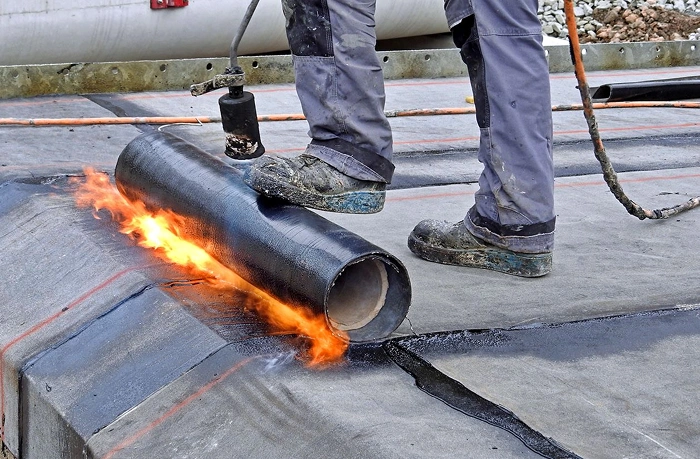 A professional roofing technician using a heat torch to install a modified bitumen roofing roll for superior waterproofing on a commercial flat roof.